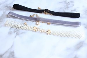 A white lace choker, a grey velvet choker with a little diamond and a black choker with a circle laying on a white marble background